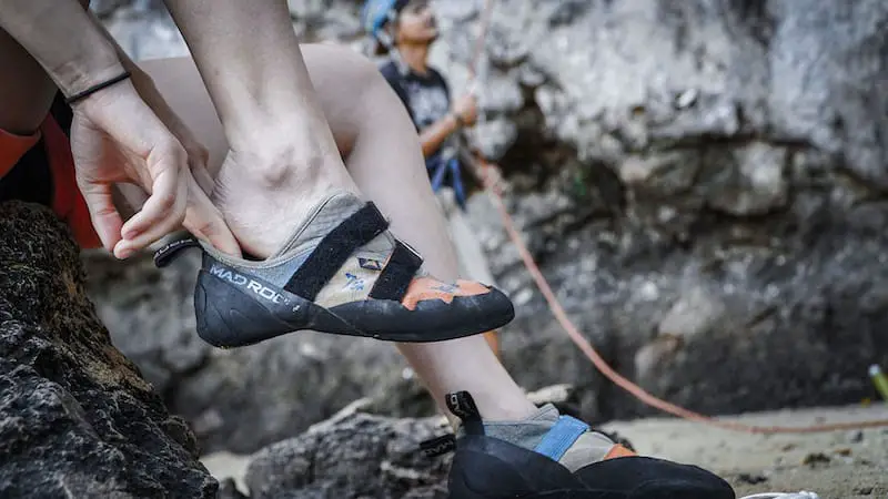man putting on mad rock climbing shoes at crag with person belay in background
